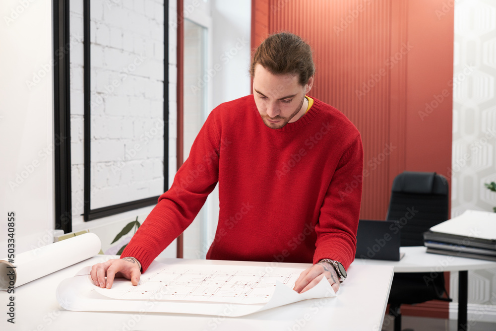 Poster young architect examining blueprint of new building at table during his work at office