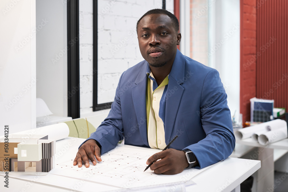 Poster portrait of african young architect looking at camera while drawing construction project at table at