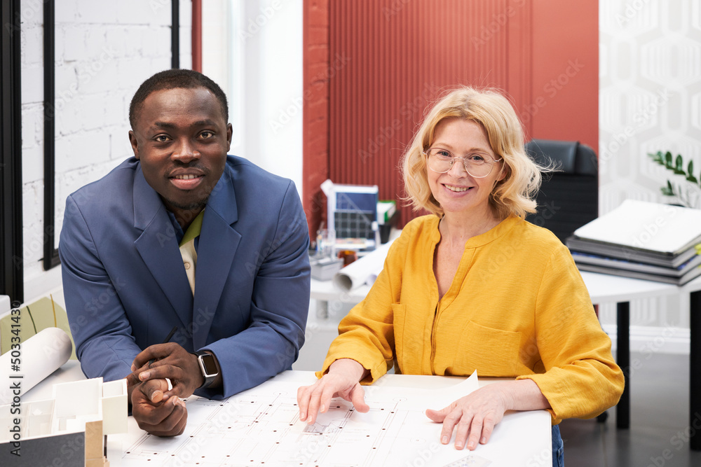 Poster Portrait of two architects smiling at camera while sitting at table and working with new project during meeting at office