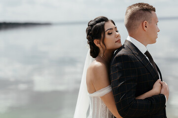 Stylish couple of elderly newlyweds stand embracing on river bank. Vertical photo