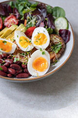 Light green salad with leaves, avocado, cherry tomatoes, cucumber slices, red beans, micro greens and medium boiled eggs on slice of toasted bread.