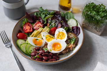 Light green salad with leaves, avocado, cherry tomatoes, cucumber slices, red beans, micro greens and medium boiled eggs on slice of toasted bread.