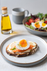 Healthy breakfast or snack with medium boiled eggs on toasted bread with cheese cream, salmon and lemon slices on grey plate. Bowl of salad and olive oil nearby