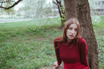 a girl in red leans on a tree and looks mysteriously into the camera