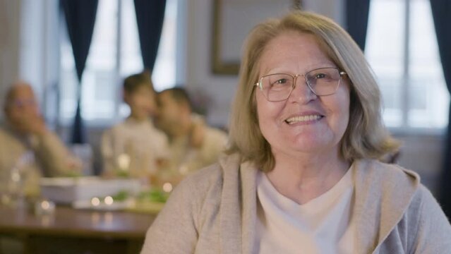 Senior blonde woman in glasses looking at camera and smiling while having nice time at family party. Blurred family image in background. Family reunion, bonding, retirement concept