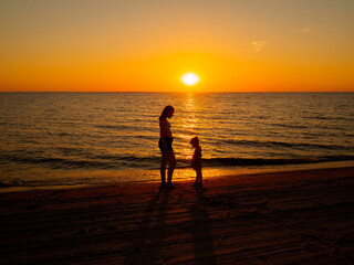 A young woman and a little girl stand opposite each other on the seashore against the backdrop of the setting sun.