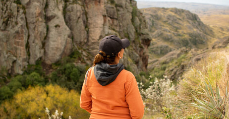 Portrait of latin woman dressed in orange with a cap and tail turned back having fun during the day of trekking in the mountain forest - Focus from the back