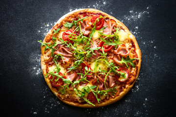 Traditional italian pizza with ham, cheese, tomatoes and arugula on black stone table. Top view.