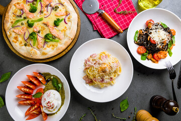 Pizza with salmon and broccoli, salad buratta with tomatoes and pesto, spaghetti carbonara with bacon, spaghetti nero black with seafood, shrimps and basil top view on grey table