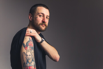 Slouchy gangster man in a black t-shirt showing to the camera tattoos on his shoulder over dark gray background. High quality photo