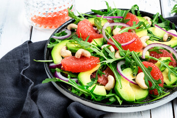 Healthy vegan salad with arugula, avocado, pink grapefruit, cashew nuts and dressing of olive oil, honey and wine vinegar. White rustic kitchen table, top view