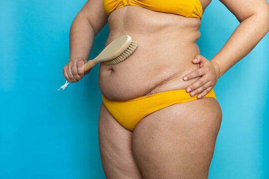 Side view of unrecognizable plus-size woman standing in yellow bra, underpants, swimsuit, brushing lymphatic drainage massage of stomach by wooden brush. Anticellulite treatment, homemade body care.