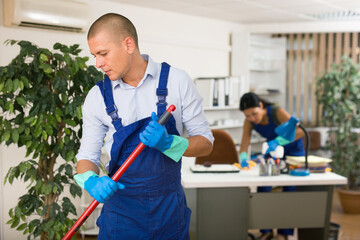 Positive male worker from cleaning service, wiping office floor with mop