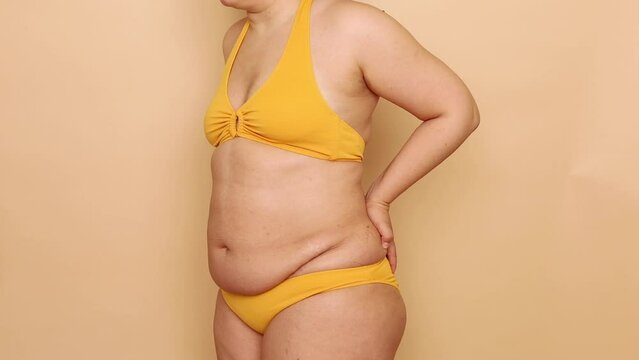 Side view of unrecognizable plump woman wearing yellow bra, bikini, showing excess naked belly, putting hands on back, posing on beige background. Body positive, obesity, weight loss, liposuction.