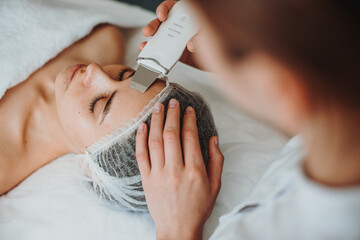 Top view of a woman receiving ultrasound cavitation facial peeling in spa salon. Happy face. Cosmetology beauty procedure. Facial skincare. - obrazy, fototapety, plakaty