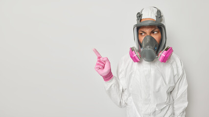 People and ecological disaster concept. Annoyed woman wears white protective suit with hood and respirator mask wears gloves takes part in special mission indicates at blank space for your promotion