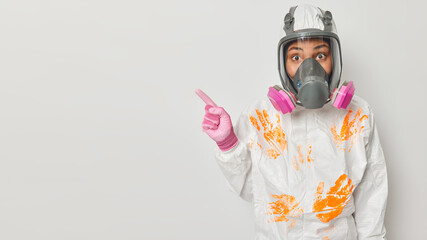 Horizontal shot of female ecologist explores surroundings measures pollution dressed in white protective suit with handprints and respirator points away on copy space asks to save planet from disaster - obrazy, fototapety, plakaty