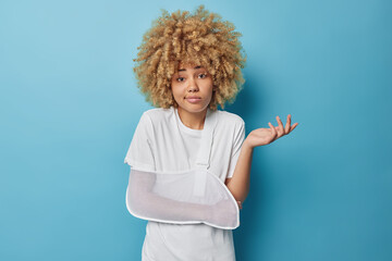 Hesitant curly haired woman has broken arm wears soft splint armband shrugs shoulders feels unaware...