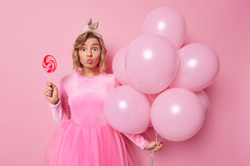 Obraz na płótnie Canvas Surprised lovely young woman keeps lips folded has hairstyle wears crown and festive dress holds lollipop and bunch of balloons comes on party isolated over pink background. Holidays concept