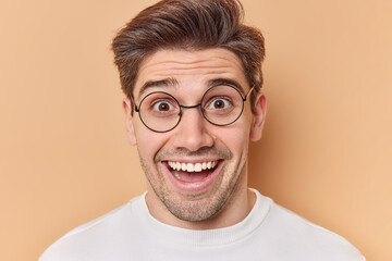 Portrait of happy surprised adult man reacts on amazing news wears round transparent eyeglasses smiles broadly dressed in casual wear isolated over brown background. Human reactions concept.