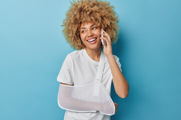 Positive curly haired young woman has broken arm talks via smartphone wears sling looks away with...