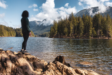 Fototapeta na wymiar Adventurous Caucasian Woman on the rocks by the water in Canadian Nature Landscape. Buntzen Lake, Anmore, Vancouver, BC, Canada. Adventure Travel Concept