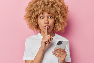 Surprised woman with curly bushy hair shows silence gesture keeps index finger over lips uses mobile phone wears casual white t shirt isolated over pink background. Don not tell anyone about this