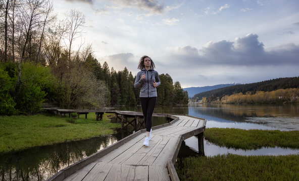 Caucasian Athletic Woman running on a Wooden Path across a swamp in Shoreline Trail, Port Moody, Greater Vancouver, British Columbia, Canada. Trail in a Modern City during a Sunny Blue Sky.