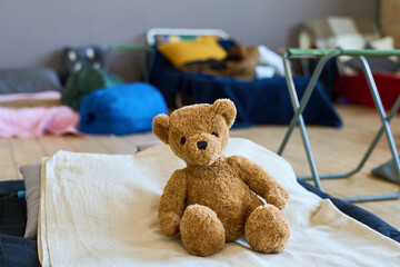 Teddybear of brown color sitting on bed done with white cotton blanket of refugee child in large...