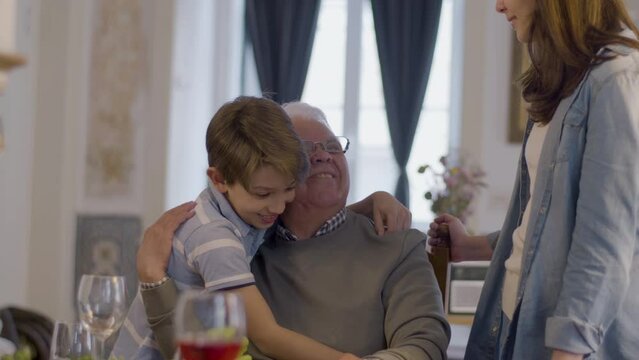 Senior white-haired man in glasses sitting at table at family party while talking to granddaughter and grandson and hugging them. Family reunion, bonding, generation gap concept