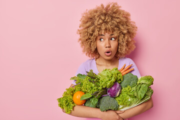 Studio shot of shocked curly haired young woman poses with groceries has amazed expression looks wondered away eats healthy vegatables isolated over pink background with blank space for promo