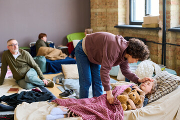 Young female refugee bending over her sick little son sleeping on couchette in spacious room prepared for homeless people