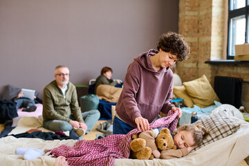 Young brunette woman covering her sleeping son with teddybear with warm blanket against other...