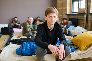 Serious boy in casualwear sitting on couchette prepared for homeless people and looking at camera against refugees talking to each other - obrazy, fototapety, plakaty