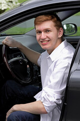 Young Male Teen Sitting in His Gray car with hand on the steering wheel.   Wearing a big smile and he has auburn red hair.