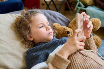 Cute blond boy in casual knitted sweater lying on couchette and scrolling in his mobile phone while...