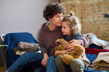 Young woman giving hug to her cute little son with brown soft teddybear while both sitting on sleeping place prepared for refugees - obrazy, fototapety, plakaty