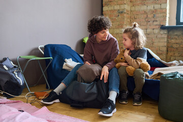 Young woman in casualwear looking at her son while sitting on couchette next to him and taking warm woolen grey sweater out of bag - obrazy, fototapety, plakaty