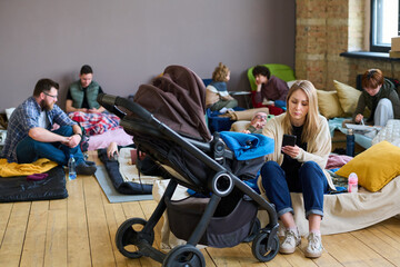 Young female with smartphone sitting on couchette by stroller with her baby against refugees and looking through online news