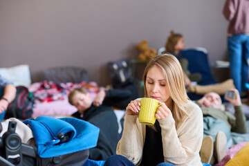 Young blond female refugee with yellow mug having hot tea while sitting on couchette by stroller with her baby against other people