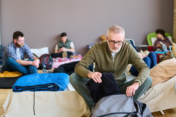 Retired man putting folded casualwear into bag while sitting on couchette against young male and...