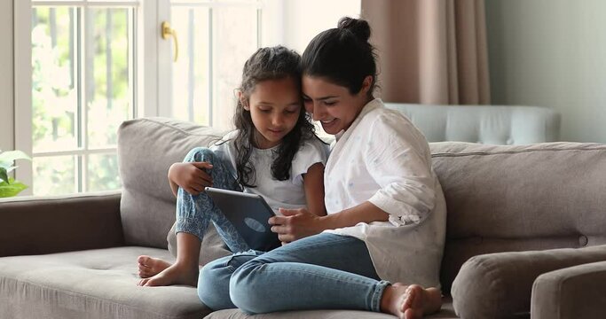 Indian mother little daughter junior schoolgirl cuddle on comfy sofa play internet video game using electronic touchpad. Two diverse age females mom child girl scroll social media pages on tablet pc