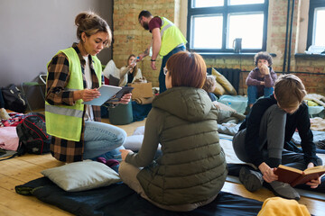 Female volunteer with mobile gadgets sitting on squats in front of refugee sitting on her sleeping...