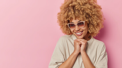 Positive young curly haired woman keeps hands under chin smiles happily focused away wears trendy sunglasses and t shirt isolated over pink background with copy space for your promotional content
