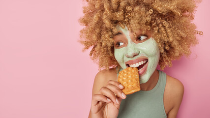 Pretty positive woman with curly bushy hair bites delicious waffle applies beauty mask on face for...