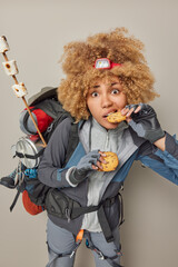 People leisure vacation concept. Hungry female camper eats cookies returns from picnic has active rest carries rucksack with necessary equipment isolated on grey wall prepared marshmallow on fire