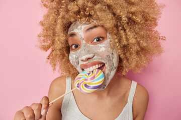 Close up portrait of cheerful curly haired woman bites caramel candy applies beauty mask on face for skin treatment wears casual t shirt isolated over pink background. Unhealthy food concept