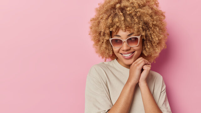 Pleased dreamy young woman with glad expression curly bushy hair keeps hands near face wears trendy sunglasses and casual t shirt isolated over pink background empty space for your promotion