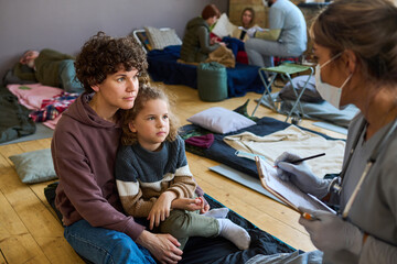 Young female refugee with little son looking at clinician in uniform and mask making notes in...