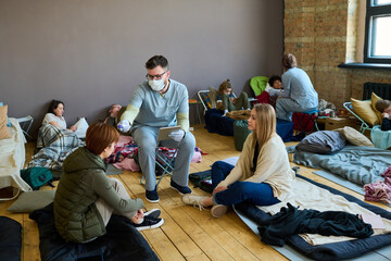 Male volunteer or healthcare worker measuring body temperature of young female refugee sitting on...